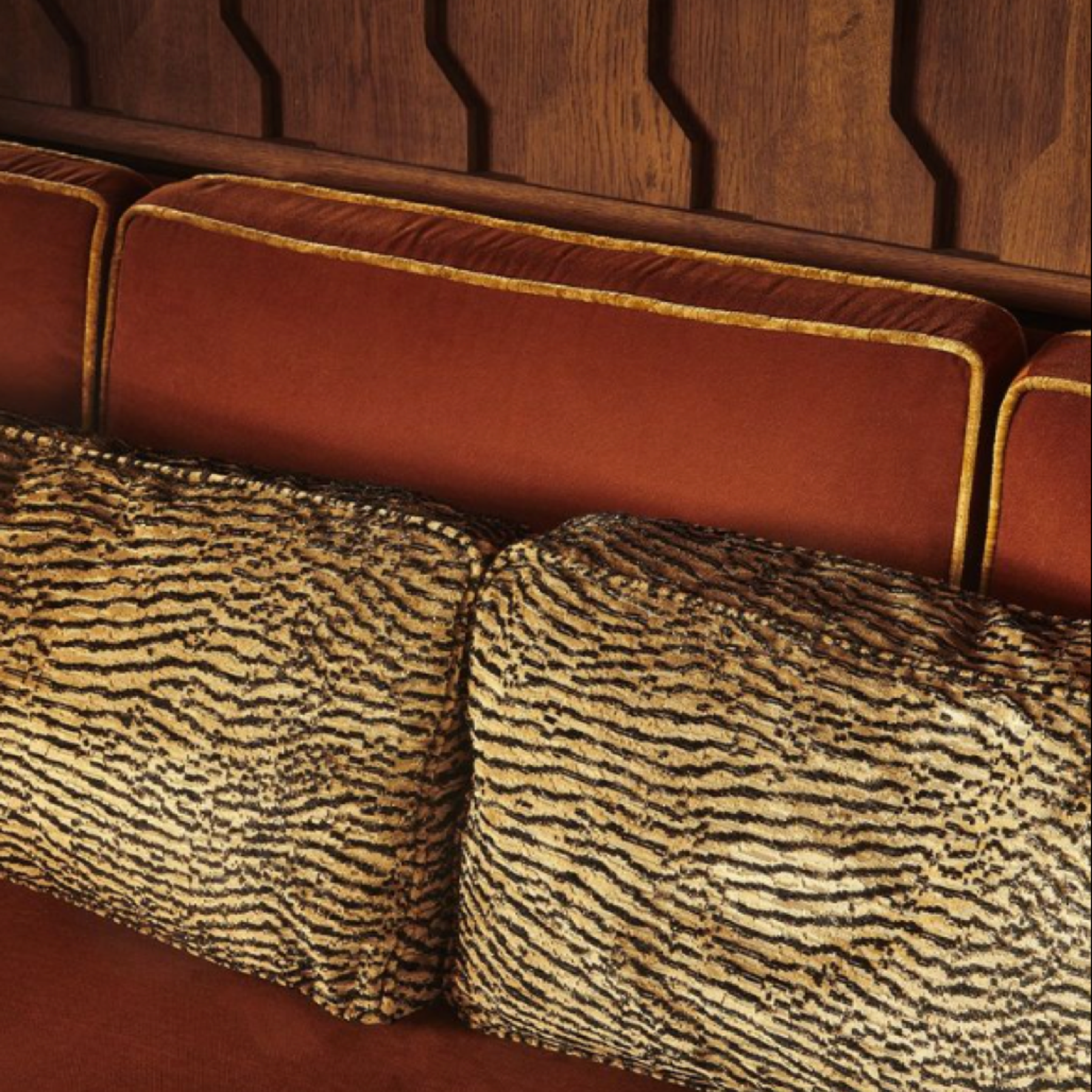 A luxurious interior featuring a rich terracotta sofa with gold piping, accented by textured animal print cushions. The backdrop showcases a sophisticated wooden panel with geometric carvings, blending modern design with African-inspired elements to create a warm and inviting space.