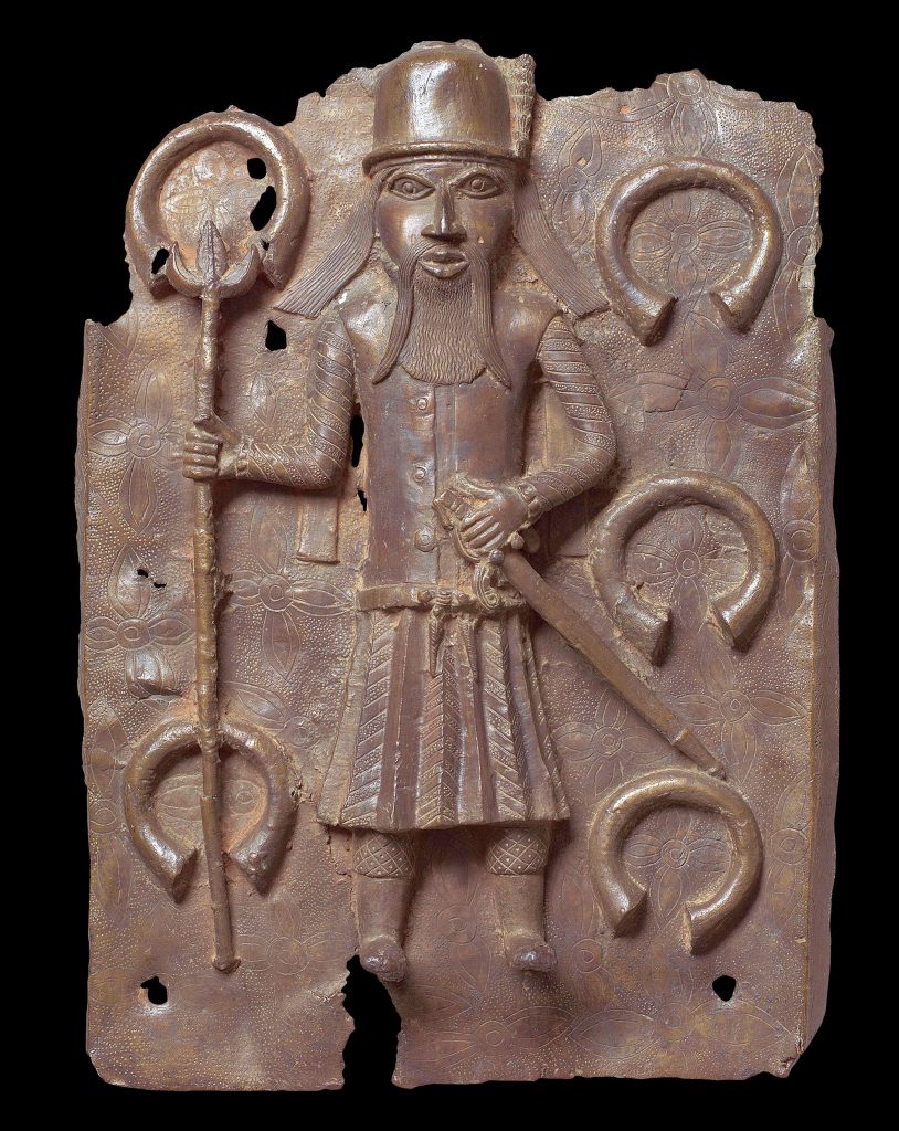Benin bronze plaque depicting a bearded Portuguese figure holding a staff, surrounded by manillas on a patterned background.