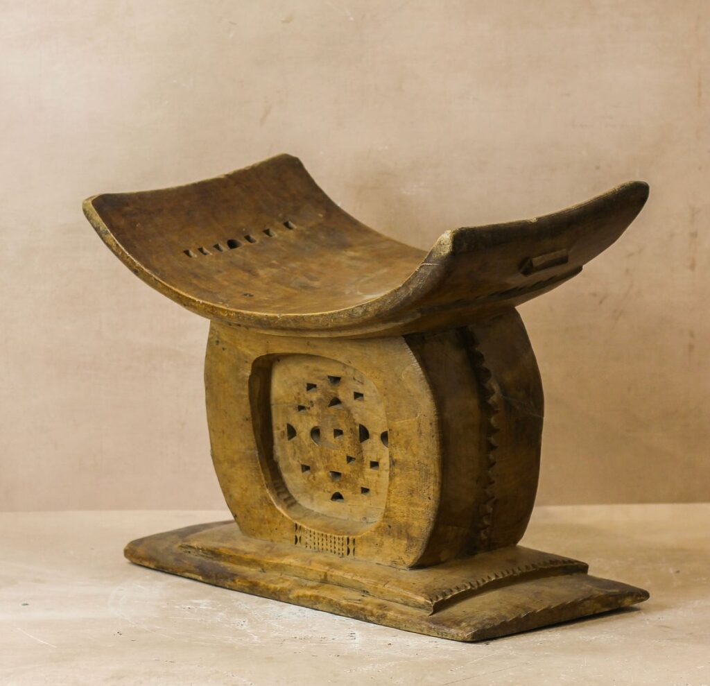 Traditional African Wooden Stool – A hand-carved wooden stool featuring a curved, saddle-like seat and a solid central base adorned with geometric perforations. The warm patina and intricate detailing suggest cultural and ceremonial significance, characteristic of traditional Akan or Ashanti design from West Africa. The piece sits on a rectangular platform, displaying a rich sense of craftsmanship and heritage.