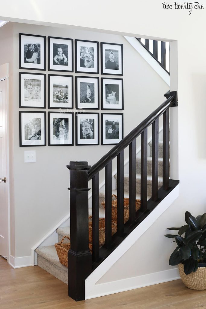 Staircase Gallery Wall with Black-and-White Photos.
A contemporary staircase with dark wood railing and light carpeted steps. The wall along the staircase is decorated with a neatly arranged grid of twelve black-and-white family photos in matching black frames. The clean, minimalist space features white walls, light wood flooring, woven baskets under the stairs, and a potted plant for a touch of greenery