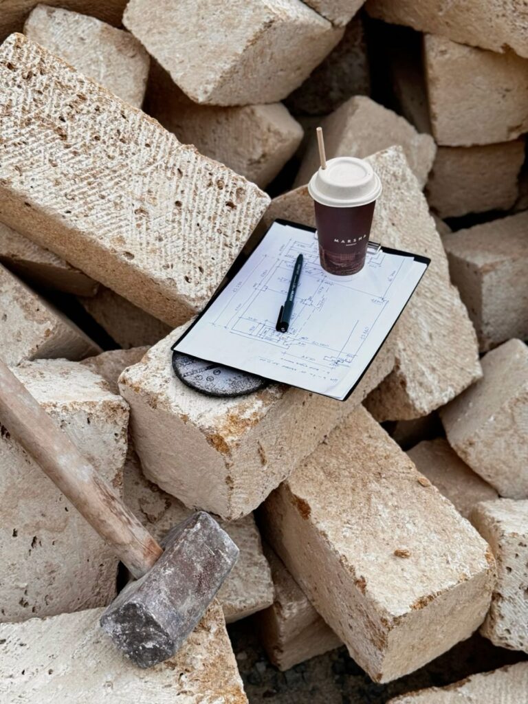 Sledgehammer leaning against stacked rough stone blocks with a coffee cup, pen, and hand-drawn plan resting on top.